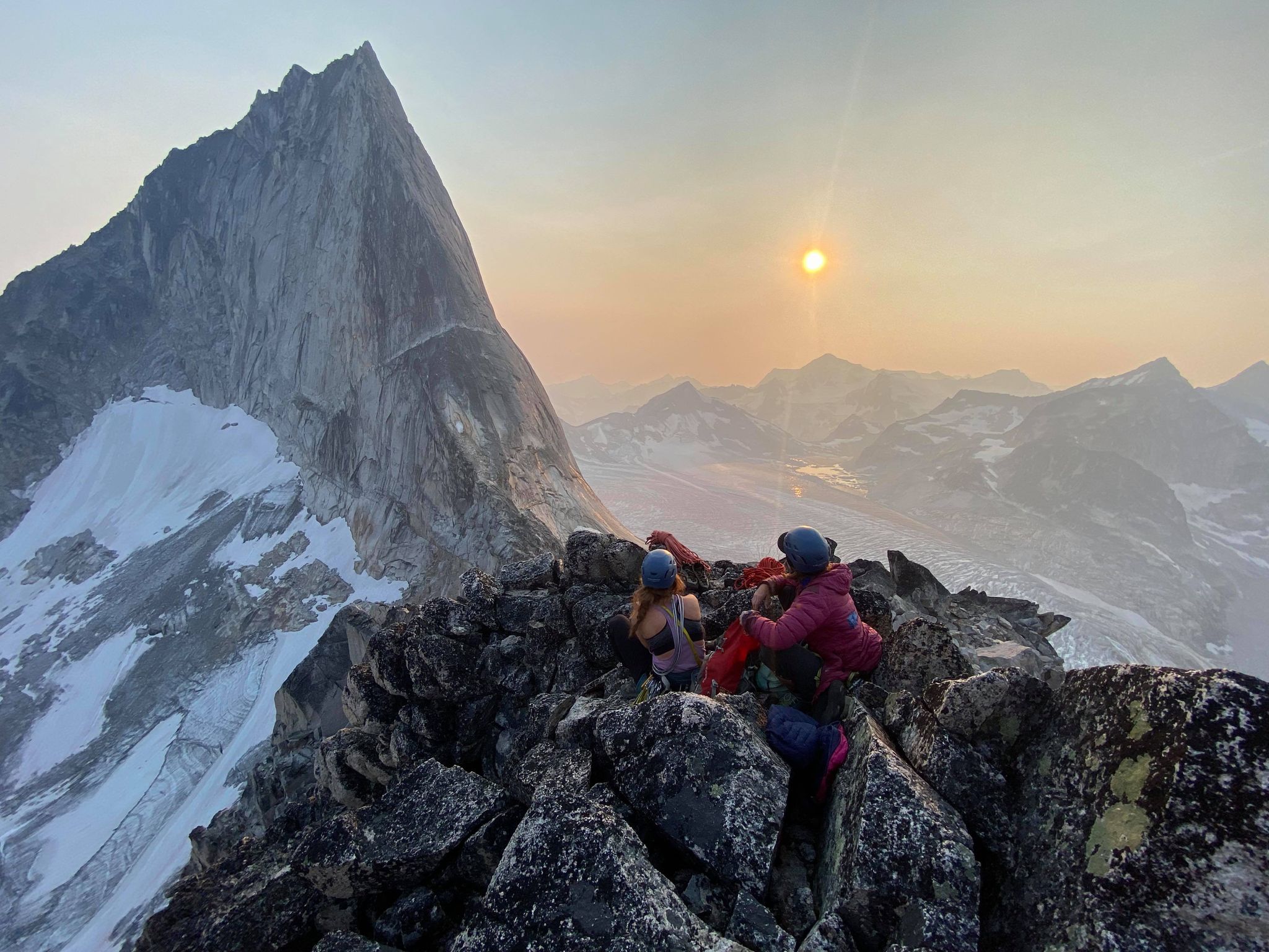 two climbers resting on a summit ridge, watching the sun set over a glacier